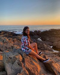 Woman sitting on rock at beach against sky during sunset