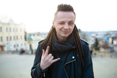 Portrait of smiling hipster gesturing horn sign while standing at terrace in city