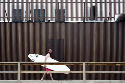 Smiling man carrying surfboard while walking by wall