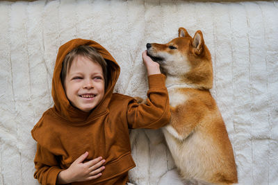 View from above on cute boy and his domestic ginger dog are lying on white cozy blanket 