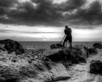 Rear view of man with tripod standing on rock against cloudy sky