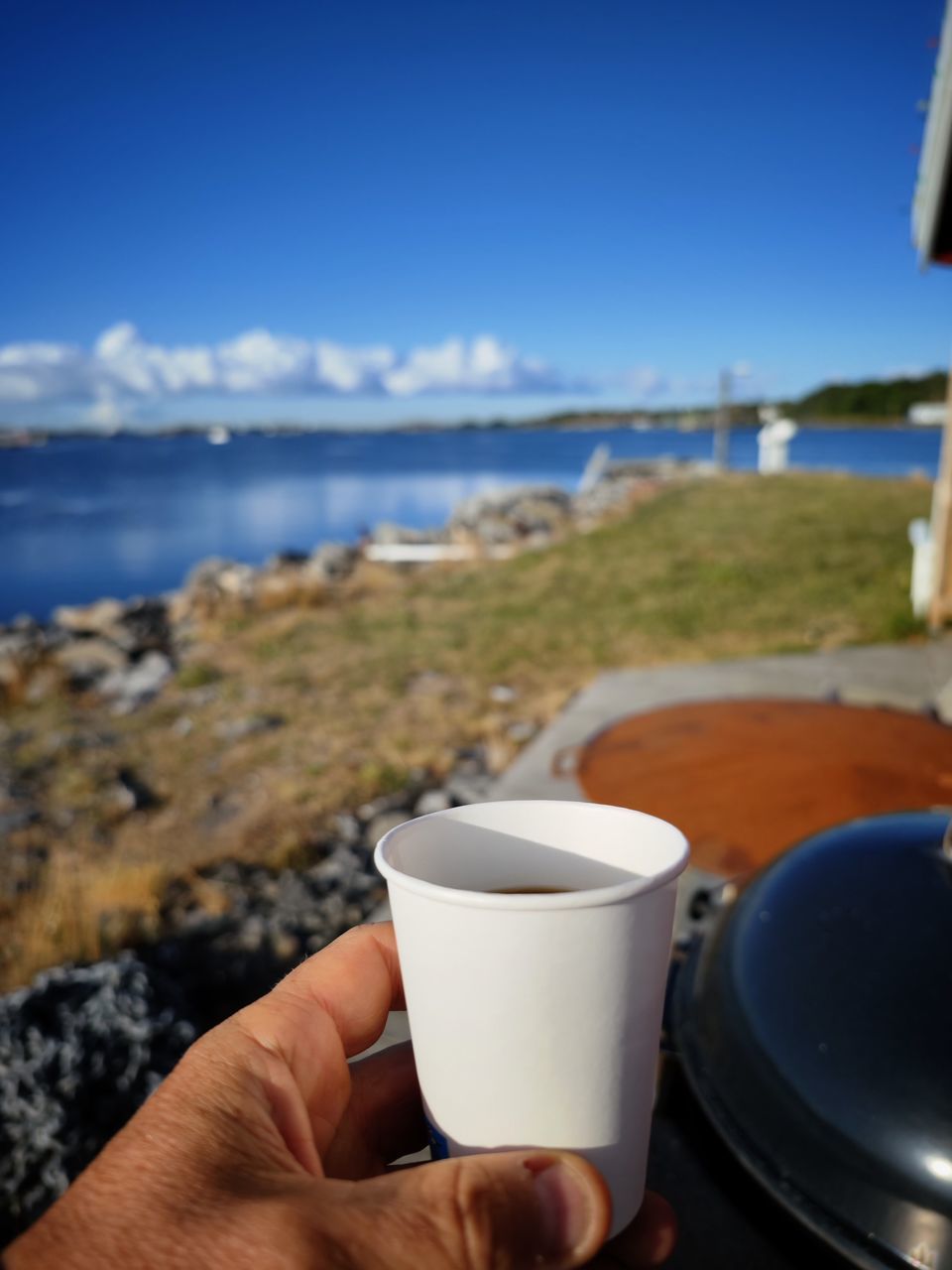water, sea, hand, drink, beach, food and drink, sky, cup, land, coffee, coffee cup, focus on foreground, nature, mug, refreshment, day, holding, leisure activity, one person, close-up, outdoors, travel destinations, vacation, personal perspective, trip, holiday, travel, adult, lifestyles, coast, horizon, blue, coastline, relaxation, scenics - nature, clear sky, food, ocean