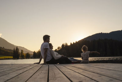 Family with little daughter sitting together at end of lakeshore jetty at sunset