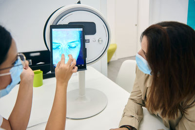 Side view of cosmetologist and customer sitting at table with facial skin diagnostics equipment while looking at tablet and discussing results of analysis in beauty clinic