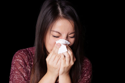 Young woman cleaning nose with facial tissue against black background