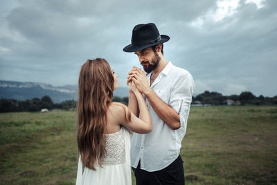 Man holding hands of woman while standing on field against cloudy sky