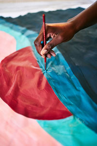 From above of crop anonymous black female with paintbrush drawing colorful picture on paper while sitting on floor at home