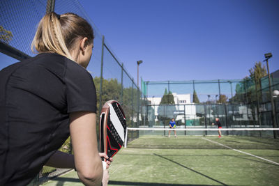 Woman looking at friends playing tennis in court against clear blue sky