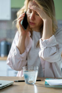 Sick woman holding sparkling water glass with aspirin pill, talking on mobile phone. vertical