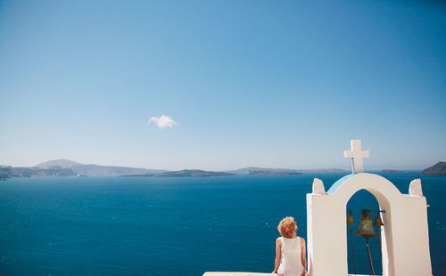 Rear view of woman sitting on church bell tower against aegean sea