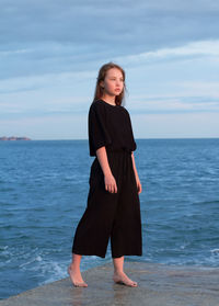 A young girl with long hair in black clothes stands on the sea pier