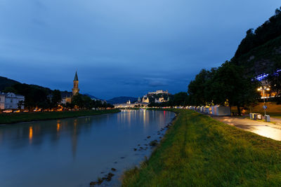 Illuminated buildings by river against sky at dusk