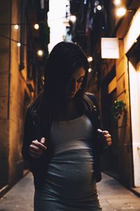 Pregnant woman standing in illuminated alley at dusk