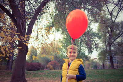 Portrait of smiling boy holding red balloon while standing in park