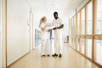 Full length of male and female nurse discussing over document in hospital corridor