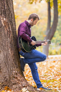 Young man playing guitar while standing by tree in park during autumn