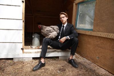 Full length of young man sitting with hen in coop