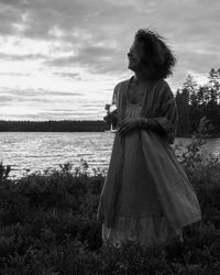 Smiling mature woman standing with drink at lakeshore against sky