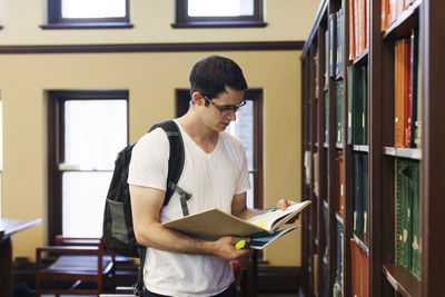 Confident man with backpack reading book while standing by shelf in library