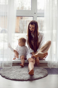 Mother in a white robe sits with a child a blonde daughter at a large window of the house person