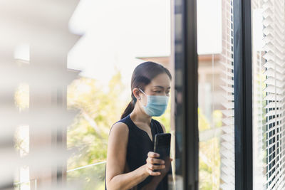 Woman in medical mask using smartphone standing by window