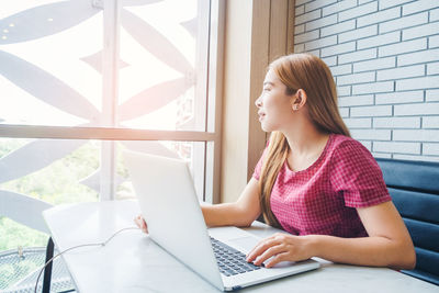 Young woman using mobile phone while sitting on window