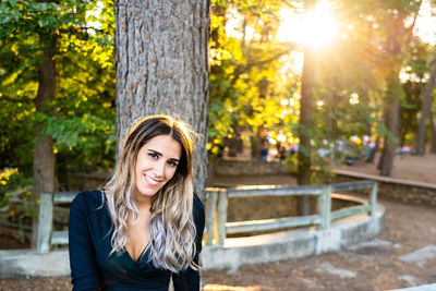 Portrait of smiling young woman against tree trunk at park