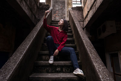 Low angle full body of young asian female traveler in casual clothes sitting on stone stairway of aged abandoned house and taking selfie while exploring streets of kaohsiung city in taiwan