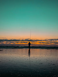 Silhouette man fishing on beach against sky during sunset