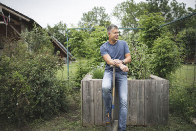 Smiling handsome man holding spade looking away while leaning on wooden raised bed at vegetable garden