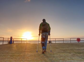 Hurt man with hooded jacket and forearm crutches looking sadly into sea water. traveler on bridge