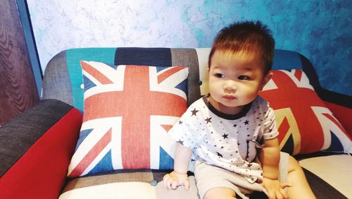 Baby boy looking away while sitting amidst british flag cushions on sofa