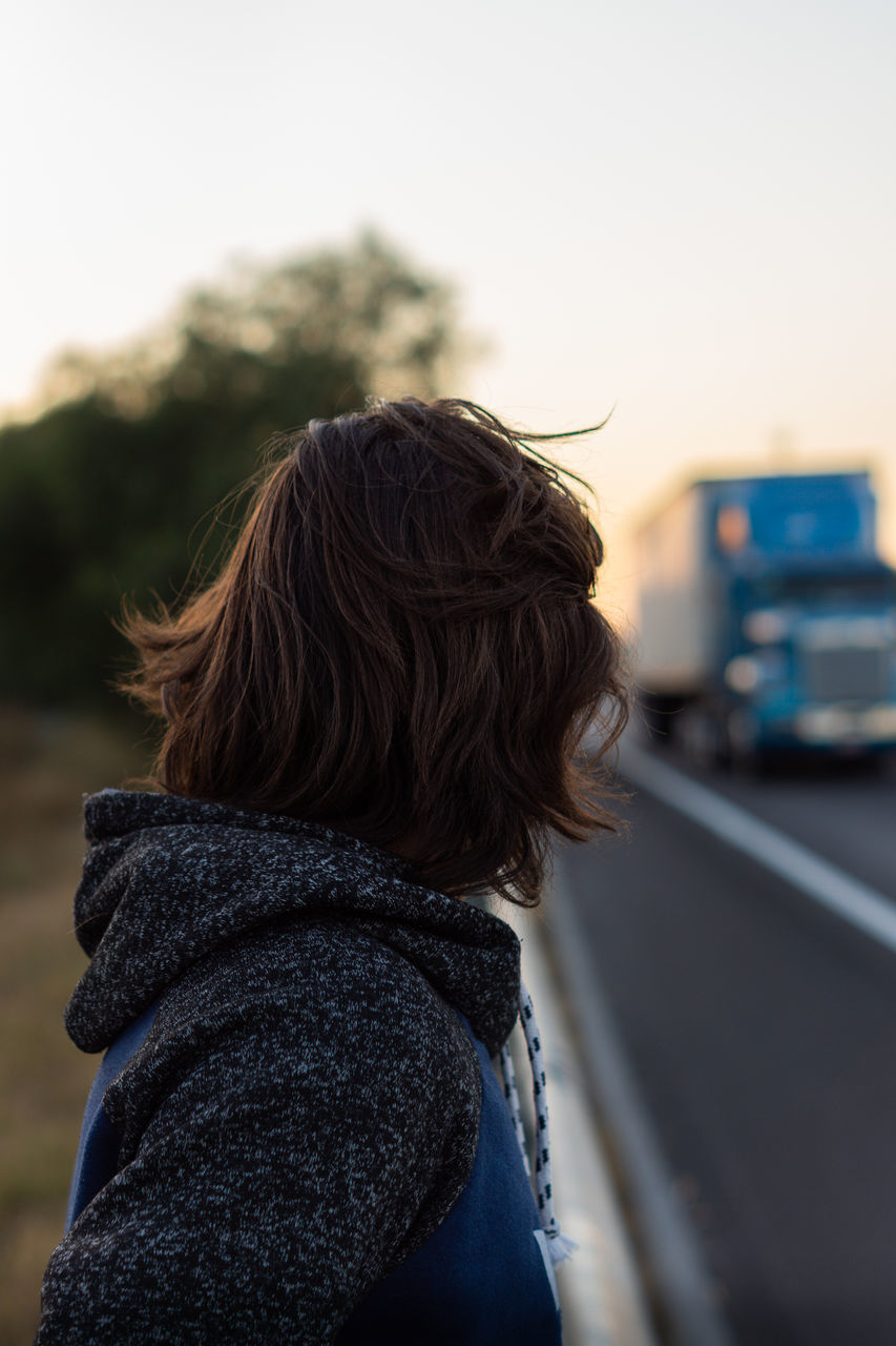 focus on foreground, real people, one person, rear view, transportation, leisure activity, headshot, lifestyles, portrait, women, mode of transportation, sky, day, clothing, adult, standing, road, motor vehicle, nature, hairstyle, warm clothing