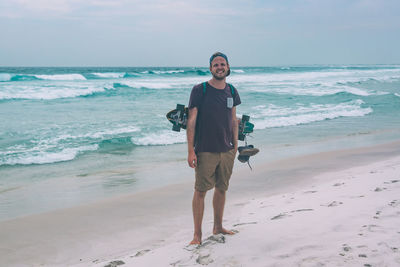 Full length portrait of young man with skateboard standing at beach