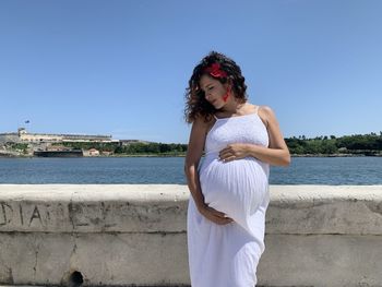 Young pregnant woman on her third trimester standing in front of water against clear sky