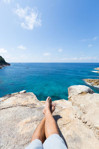 Low section of man relaxing on rock by sea against sky during sunny day