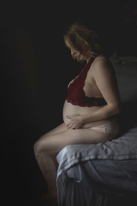 Side view of pregnant woman in lingerie touching belly while sitting on bed at home