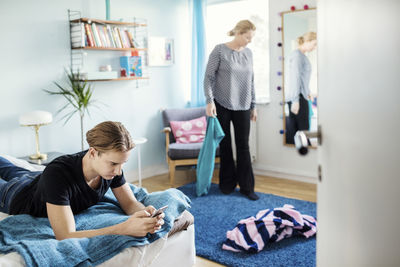 Man using smart phone while lying on bed with mother holding clothes at new house