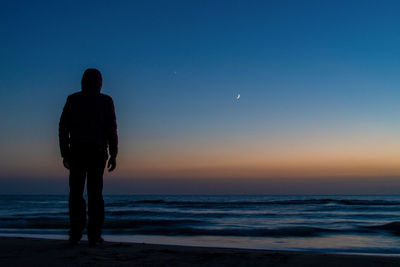 Rear view of man standing at beach against clear sky during sunset