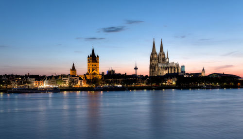 Illuminated buildings and cologne cathedral by rhine river in city at dusk