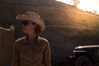 Young woman wearing sunglasses and hat looking away while standing against mountain during sunset