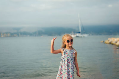 Little girl with long blonde hair stands with her back on the seashore in summer