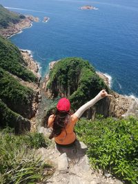Rear view of woman gesturing sitting on rock while looking at sea