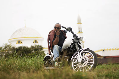 Surface level portrait of man sitting on motorcycle against mosque