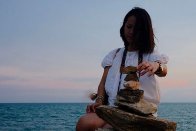 Low angle view of woman stacking rocks by sea against sky