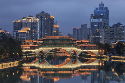 Illuminated bridge over river by buildings against sky in city