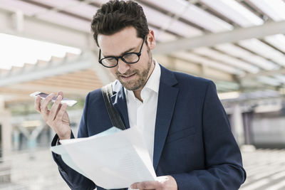 Businessman standing on parking level, reading documents, speaking notes on smartphone