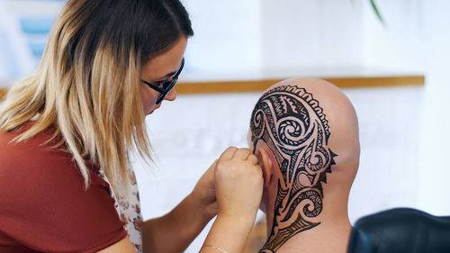 A girl, tattoo master, mehendi artist makes drawing of henna tattoo on scalp of bald caucasian man