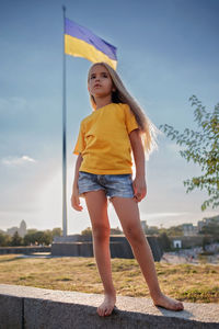 Girl in yellow polo and blue shorts near the national flag of ukraine. pray for peace and victory