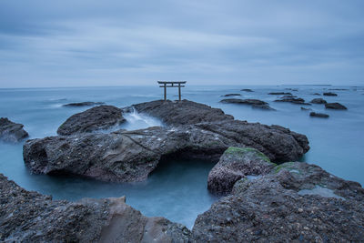 Torii gate on rock in sea against cloudy sky